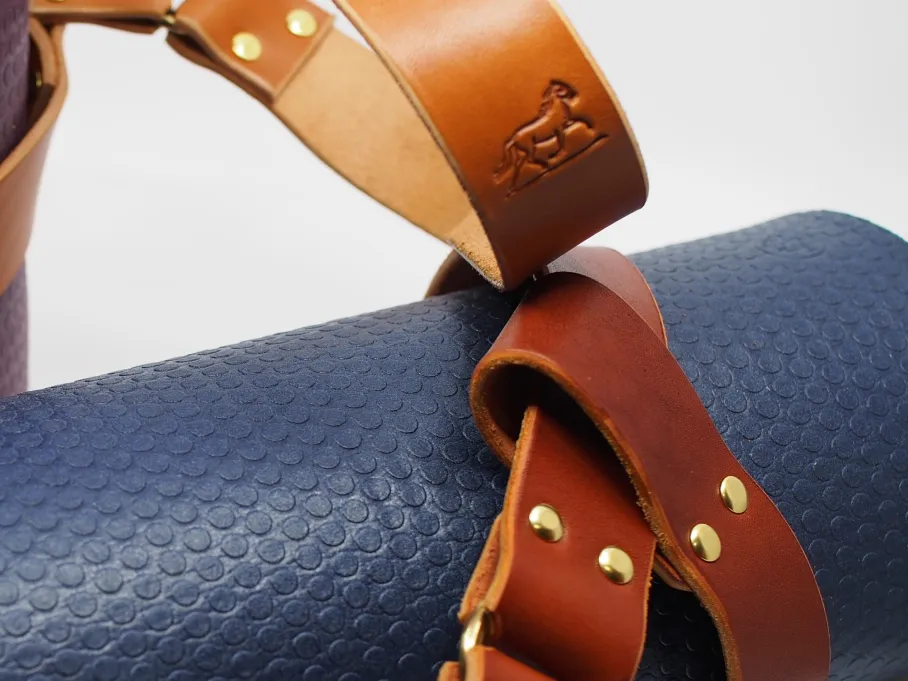 shiny brass rivets and a brass ring hold a looped chestnut strap around a navy blue yoga mat; a purple yoga mat in the background is held by a tan strap